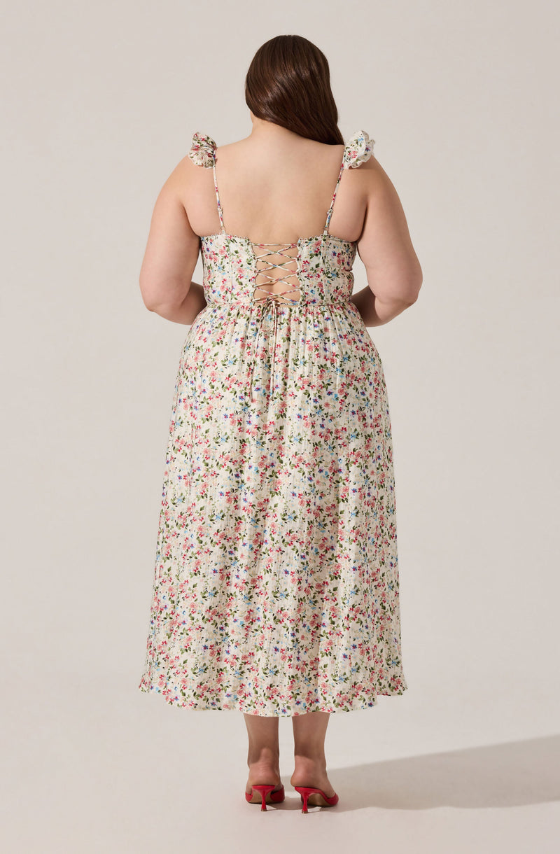 A woman is wearing a sleeveless, ankle-length dress with a floral pattern and a fitted bodice. The dress features ruffled shoulder straps and a lace-up back design. She is also wearing red high-heeled shoes.