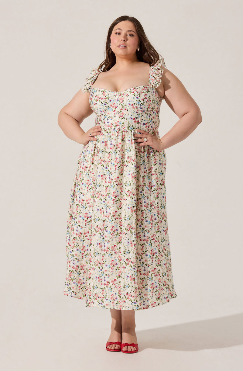 A woman is wearing a sleeveless, long floral dress with ruffled shoulder straps. The dress has a fitted bodice and a slightly gathered waist, flowing down to her ankles. She is also wearing red open-toe sandals.