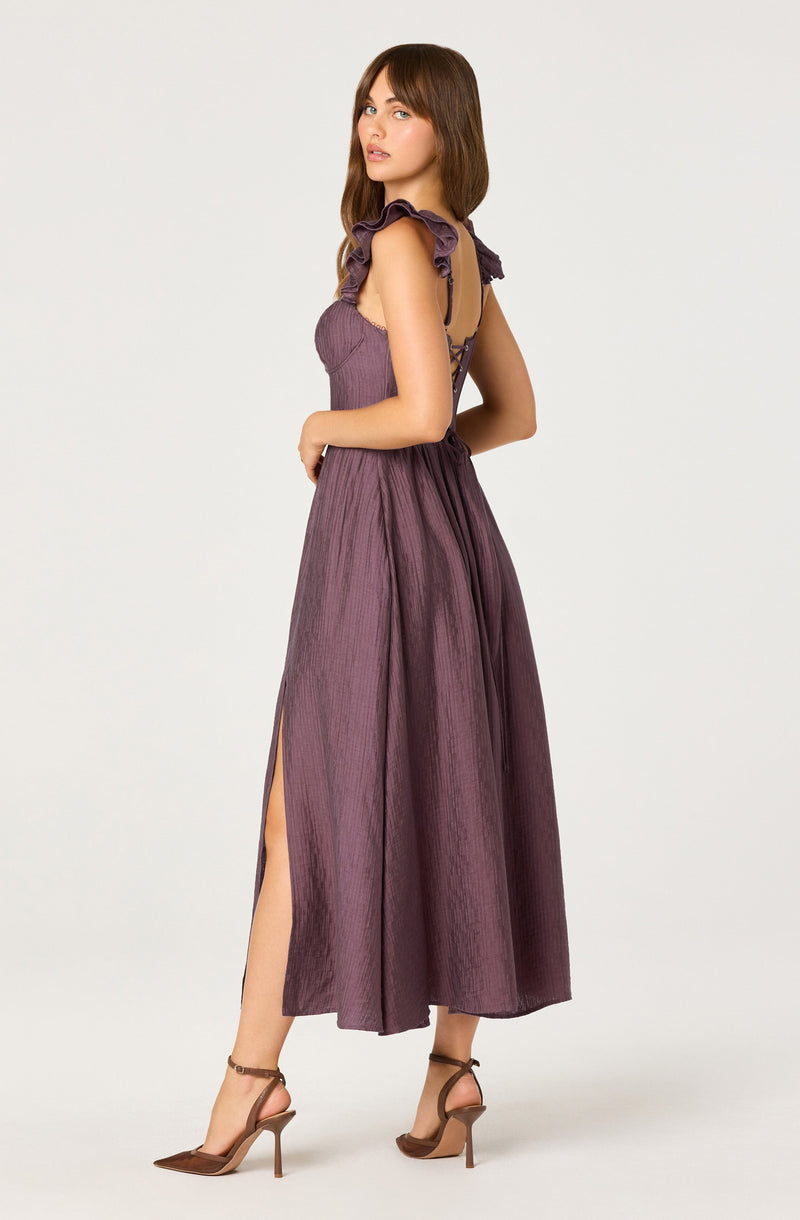 A woman is wearing a sleeveless, purple maxi dress with ruffled shoulder straps and a fitted bodice. The dress features a high slit on the side, revealing her leg. She is also wearing brown high-heeled sandals.
