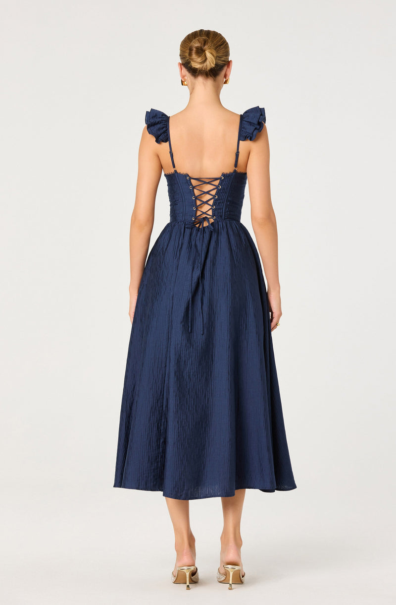 A woman is wearing a navy blue dress featuring a fitted, textured bodice with ruffled shoulder straps. The back of the dress has a corset-style lace-up detail, creating a crisscross pattern. The skirt is long, flowing, and has a shiny, crinkled fabric appearance. She is also wearing nude-colored high heels with a buckle detail.
