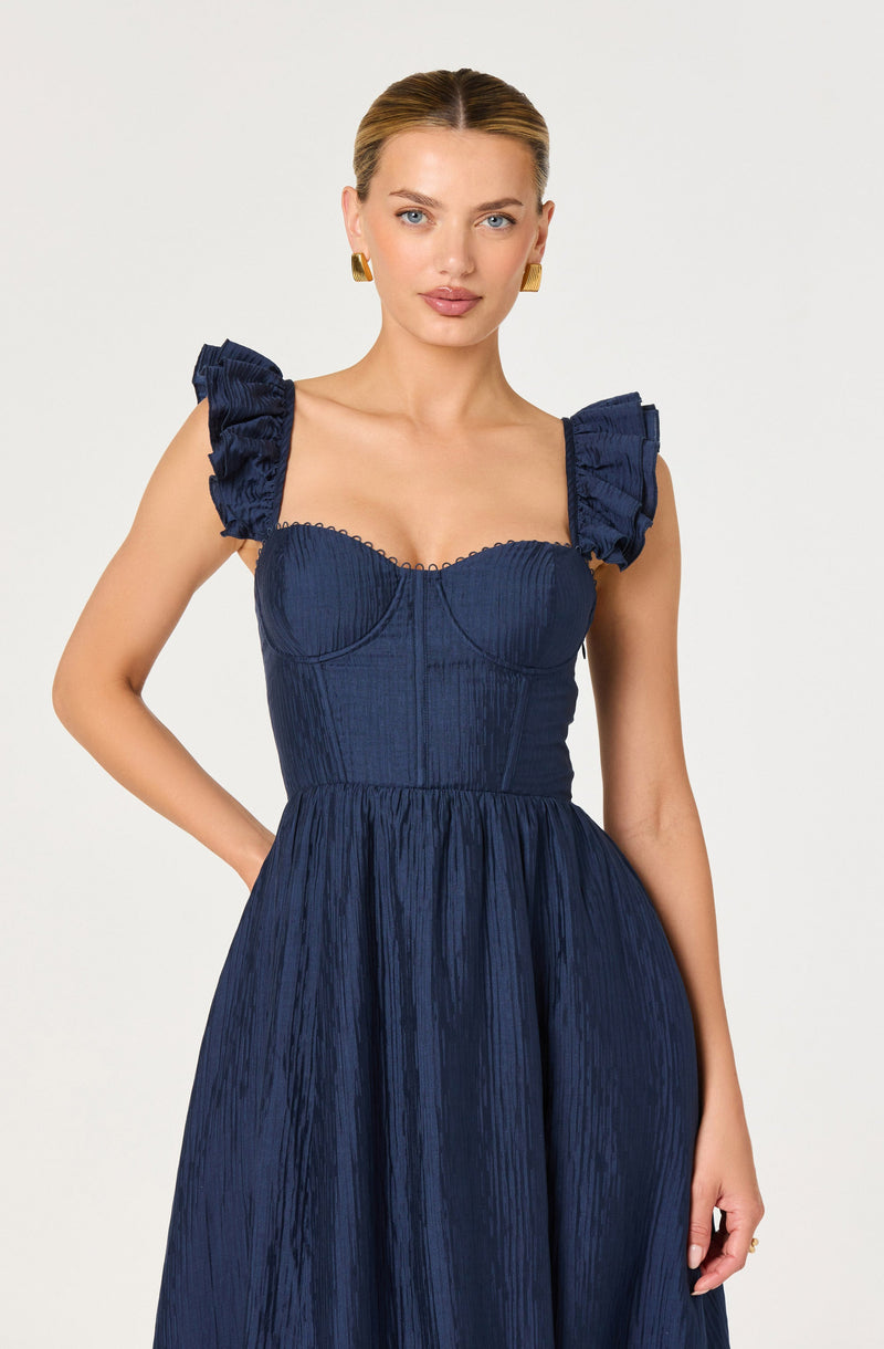 A woman is wearing a dark blue dress with puffed, ruffled shoulder straps. The dress features a fitted, structured bodice with visible vertical seams and a textured, pleated fabric. The skirt is full and gathered, flowing from the waist down in the same textured material. The overall style of the dress is elegant and fashionable.