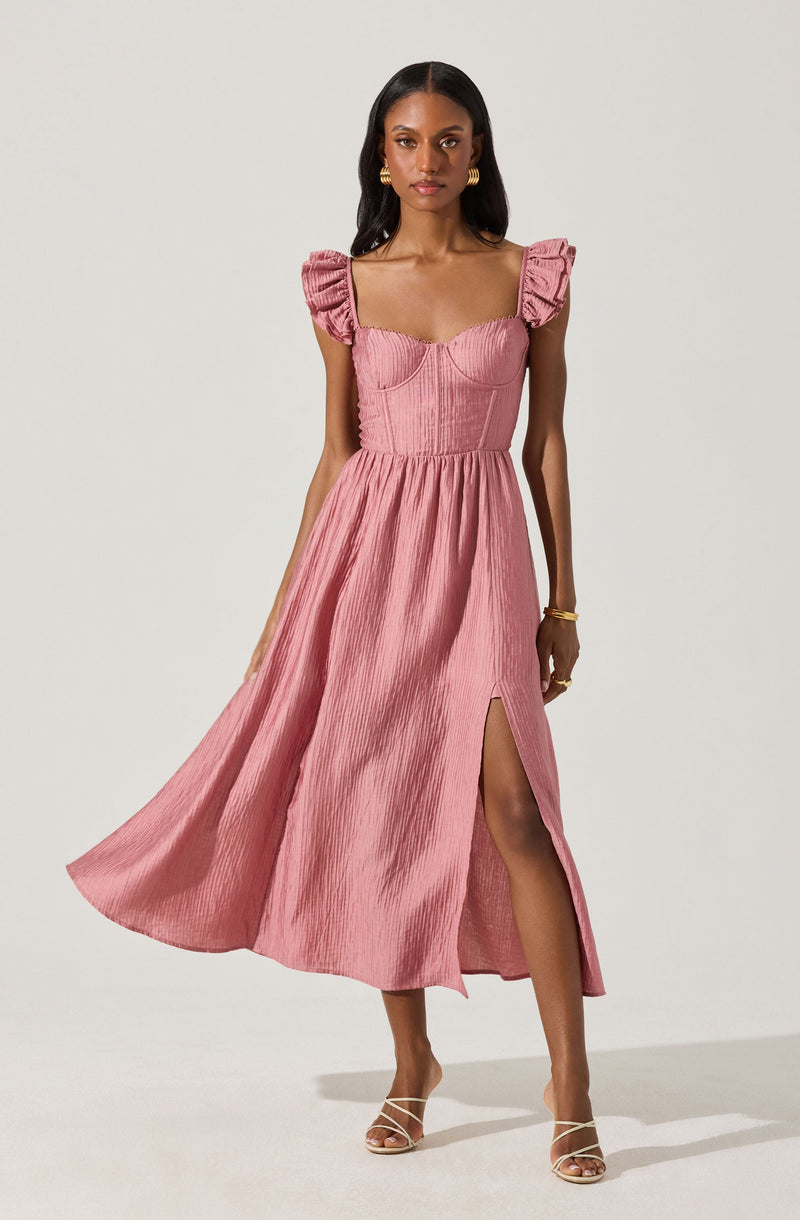 A woman is wearing a pink maxi dress made of a textured, shiny fabric. The dress features puffed cap sleeves with gathered detailing, a fitted bodice with visible seam lines, and a high slit on the left side that reveals her leg. She is accessorized with gold hoop earrings, a gold bracelet, and strappy high-heeled sandals.
