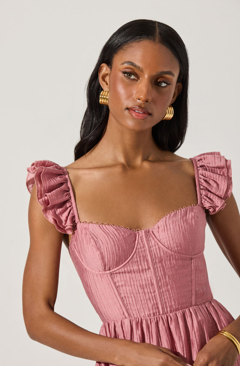 A woman is wearing a pink, textured, gathered dress with puffed short sleeves featuring ruffle details. The dress has a fitted bodice with visible seam lines and a gathered waistline. She accessorizes with large, gold hoop earrings and a gold bracelet.