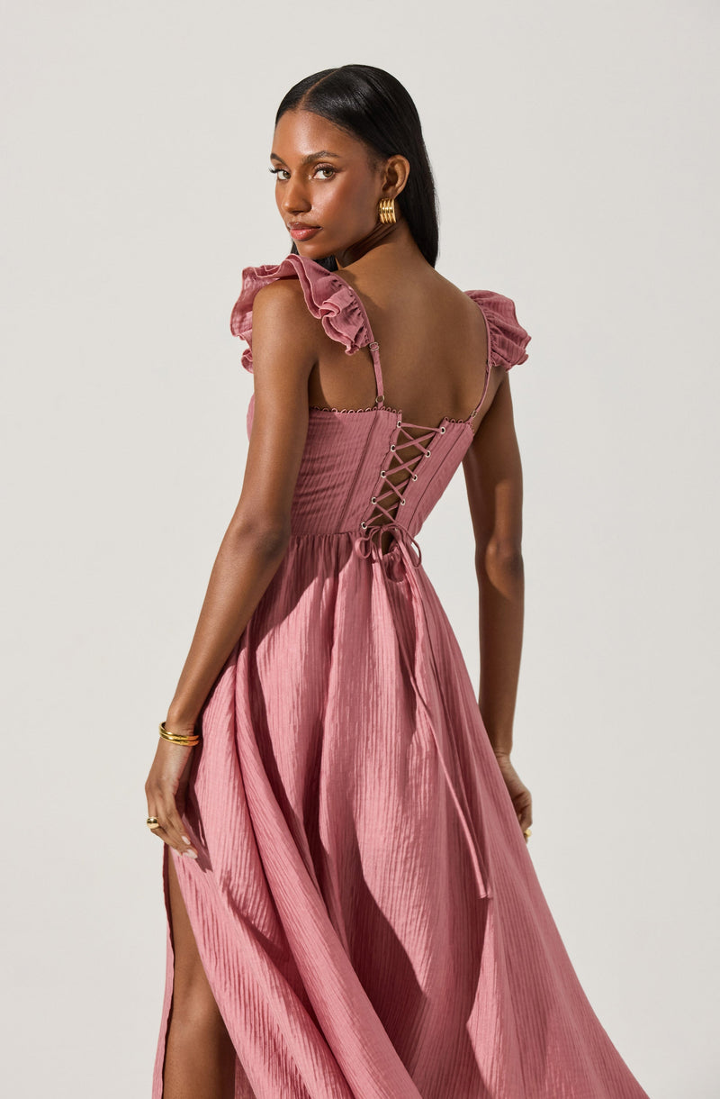 A woman is wearing a pink dress with a fitted bodice that features a lace-up back detail. The dress has ruffled, shoulder straps and a flowy, textured skirt with a slit on one side. She accessorizes with gold hoop earrings and holds her hand gracefully at her side.
