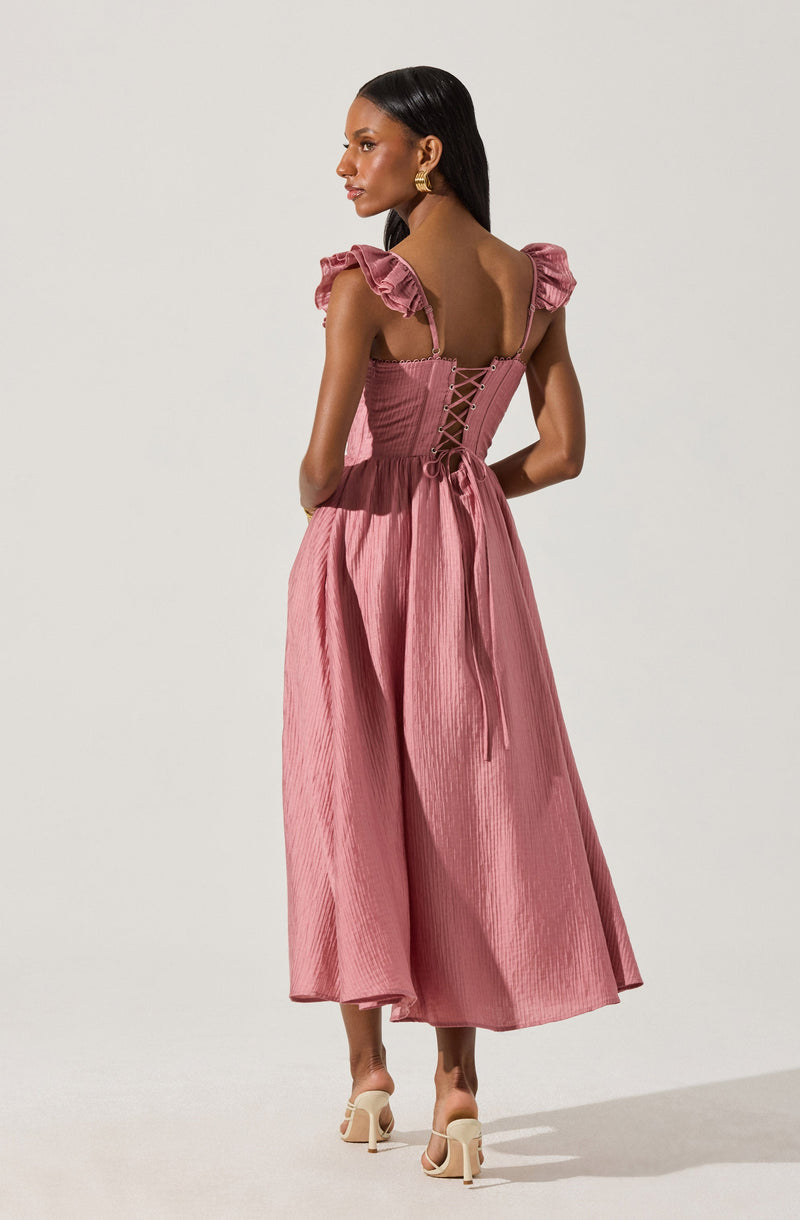 A woman is wearing a pink dress with ruffled shoulder straps and a fitted bodice that features a lace-up detail at the back. The dress has a textured, pleated fabric and a full, flowing skirt that reaches her mid-calf. She is also wearing beige high-heeled sandals and gold earrings.