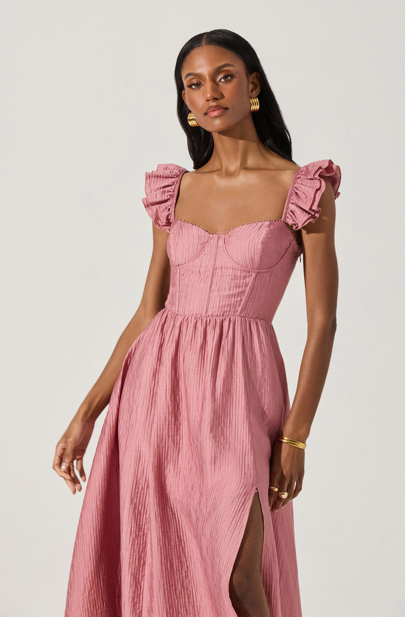 A woman is wearing a pink dress with textured, pleated fabric. The dress features short, ruffled puff sleeves and a fitted bodice with visible seam details. The skirt of the dress is long, flowing, and has a high slit on the left side.