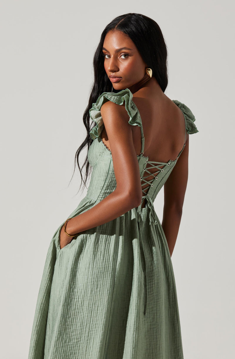 A woman is wearing a light green, pleated dress with ruffled cap sleeves. The dress features a fitted bodice with a lace-up back, tying in a bow at the waist. She has long, dark hair and is accessorized with gold hoop earrings and a gold bracelet.