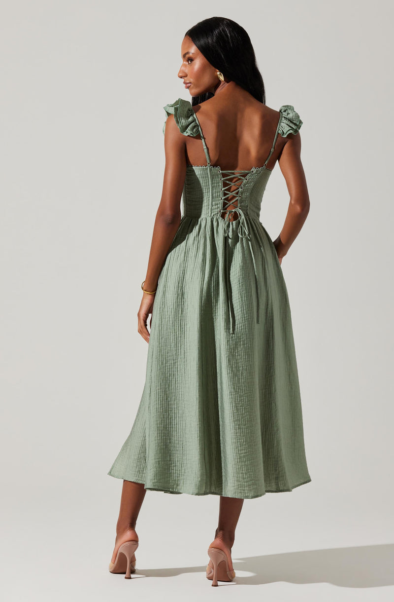 A woman is wearing a light green midi dress with a textured, crinkled fabric. The dress features ruffled shoulder straps, a fitted bodice with crisscross lace-up detailing at the back, and a tie around the waist. She is also wearing beige high-heeled shoes and gold jewelry, including earrings and a bracelet.