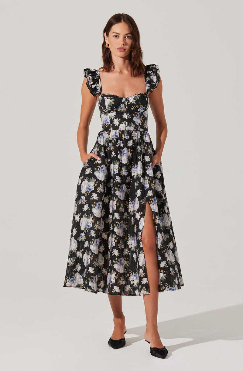 A woman is wearing a black dress with a floral pattern featuring white and light purple flowers. The dress has ruffled short sleeves, a fitted bodice, and a flowing skirt with a slit on one side.