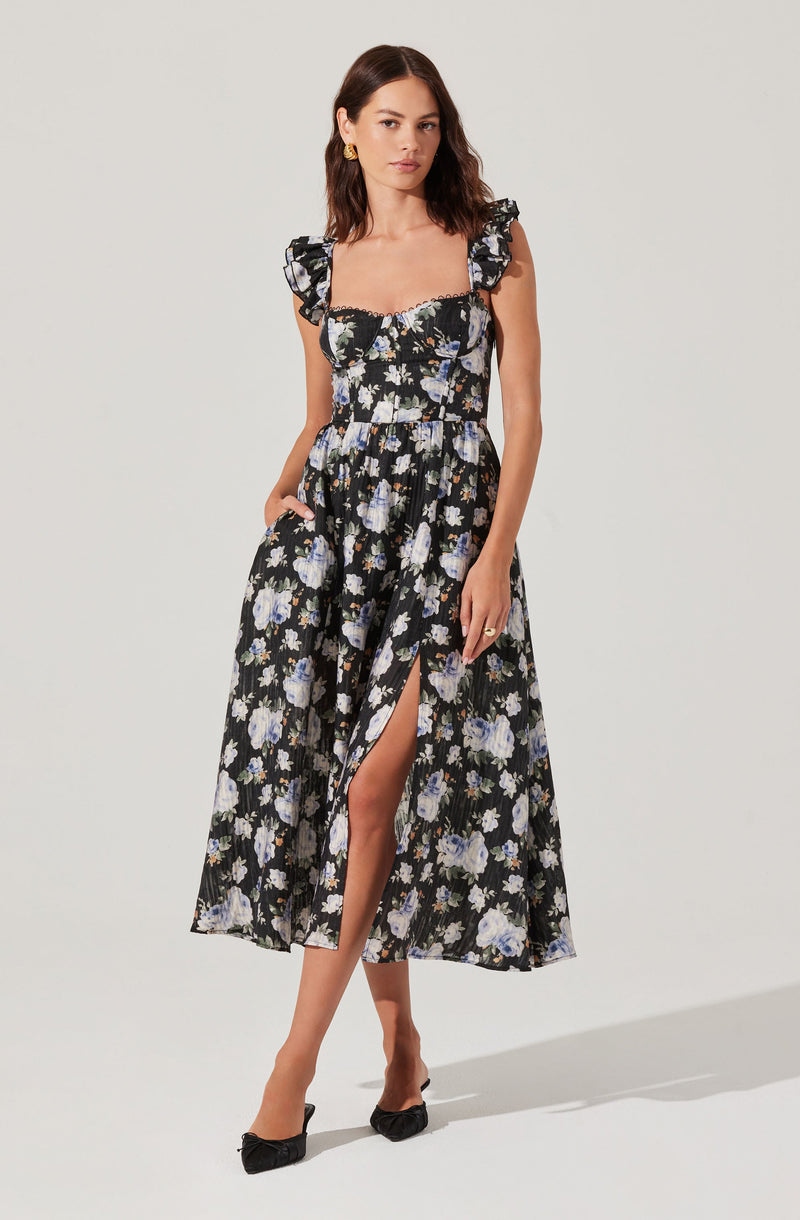 A woman is wearing a black midi dress with a floral pattern in shades of white, blue, and purple. The dress features ruffled shoulder straps, a fitted bodice with a sweetheart neckline, and a slit on the left side revealing her leg. She is accessorizing with gold earrings, rings, and black shoes with bows.