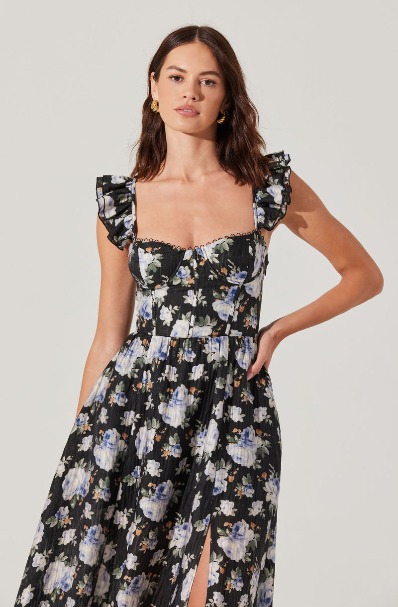 A woman is wearing a black dress with a floral pattern of white, purple, and green flowers. The dress features ruffled short sleeves, a fitted bodice with vertical seams, and a gathered skirt with a side slit. The fabric appears to be lightweight with a textured, pleated finish.