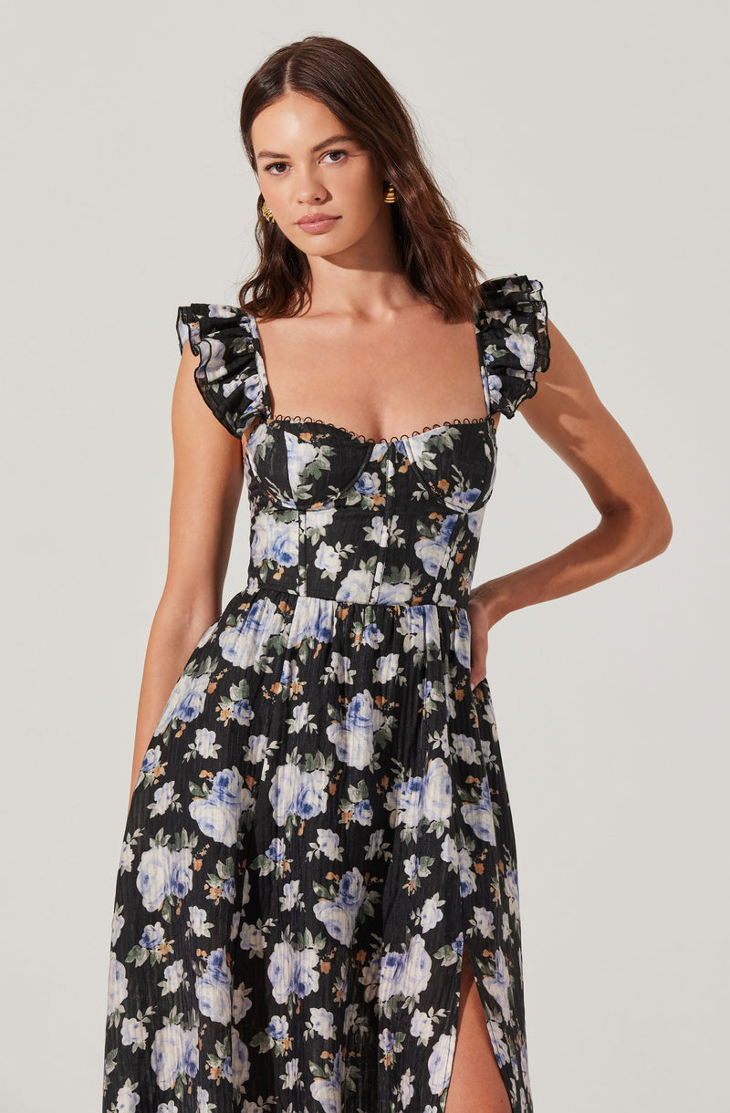 A woman is wearing a sleeveless black dress with a floral pattern of white, blue, and purple flowers, accented by ruffled shoulder straps and a slit on the left side.