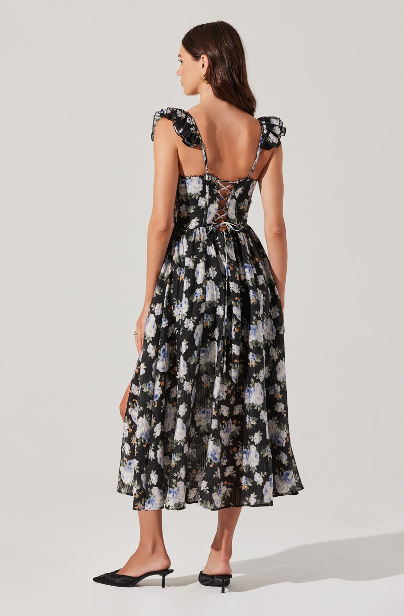 The dress features a black background with a floral pattern of white, blue, and purple flowers. It has ruffled shoulder straps, a fitted bodice with a lace-up back, and a flowy, midi-length skirt with pleats. The material appears lightweight and semi-sheer. The woman is also wearing black heeled mules and gold jewelry.