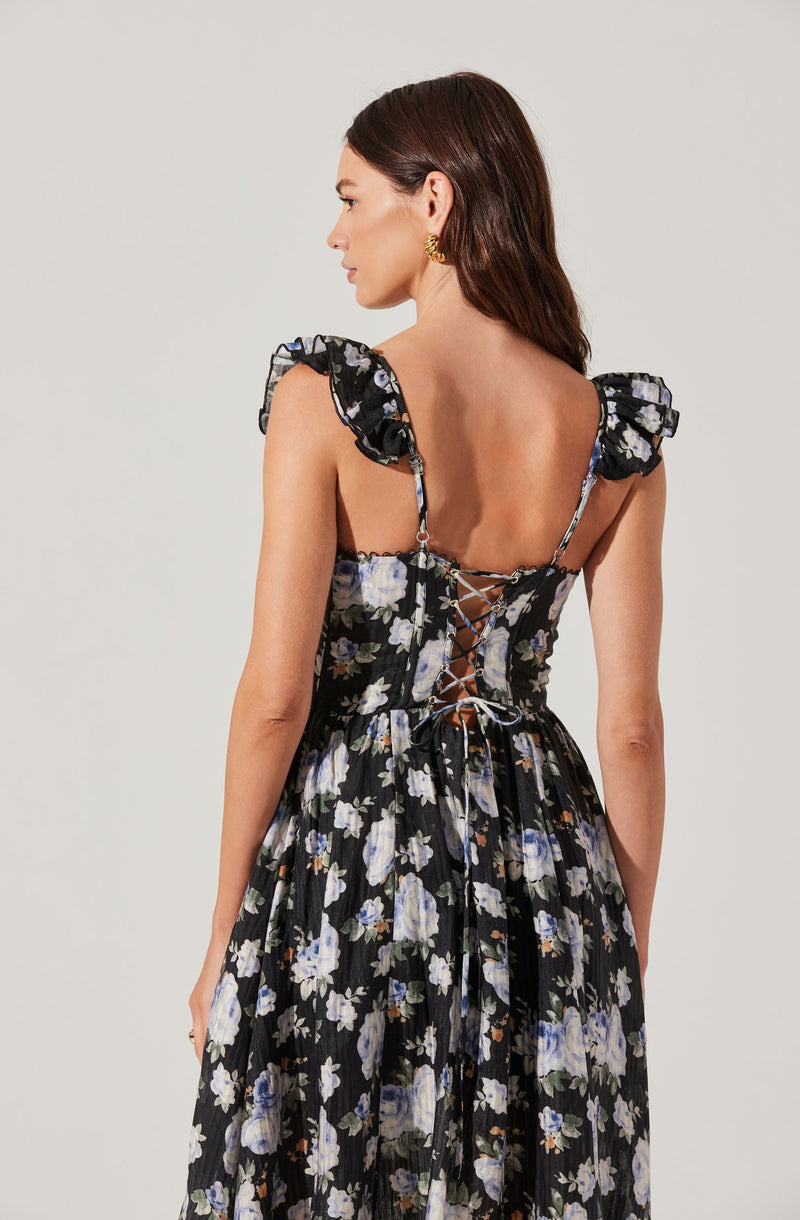 A woman is wearing a black dress with a floral pattern featuring white and purple flowers. The dress has ruffled shoulder straps and a fitted bodice with a lace-up back design.