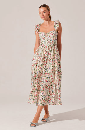 A woman is wearing a long, sleeveless dress with ruffled shoulder straps and a fitted bodice. The dress features a colorful floral pattern on a cream background, with small, multicolored flowers and green leaves. She has paired it with open-toed heels that have bows on the front.