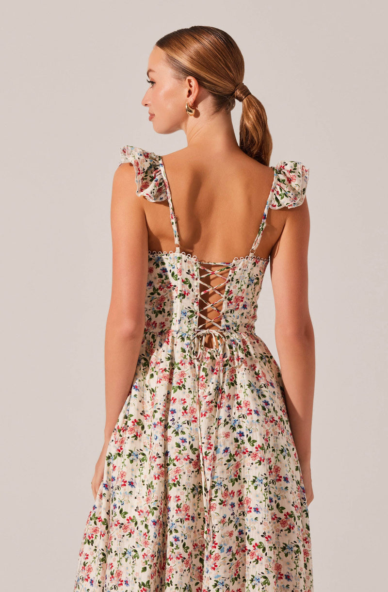 A woman is wearing a dress with a floral pattern on a light background. The dress features ruffled shoulder straps and a lace-up back with crisscrossing ties. The fabric appears lightweight with small, colorful flowers scattered throughout.