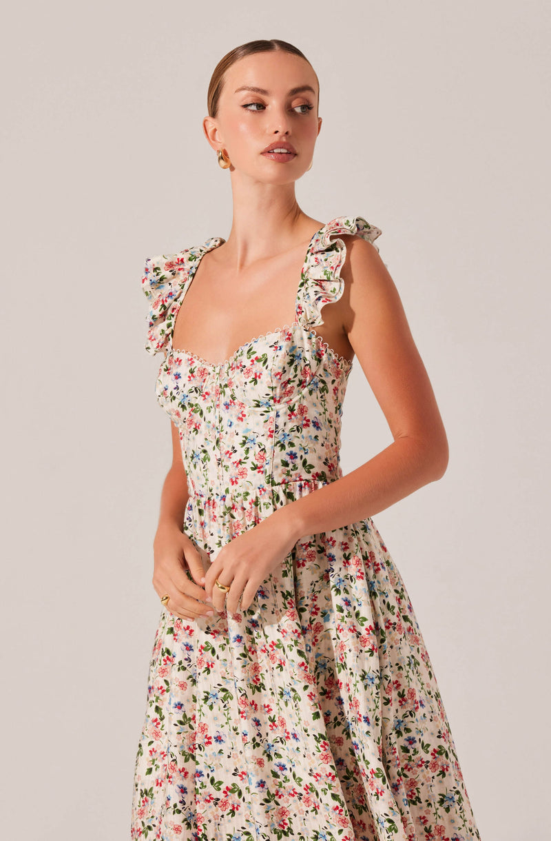 A woman is wearing a sleeveless, floral-printed dress with ruffled shoulder straps. The dress features a fitted bodice with a sweetheart neckline and gathered fabric at the waist, flowing into a long skirt. The floral pattern includes small, colorful flowers on a light background. She has gold hoop earrings and rings on her fingers, and her hair is styled in a sleek, pulled-back look.