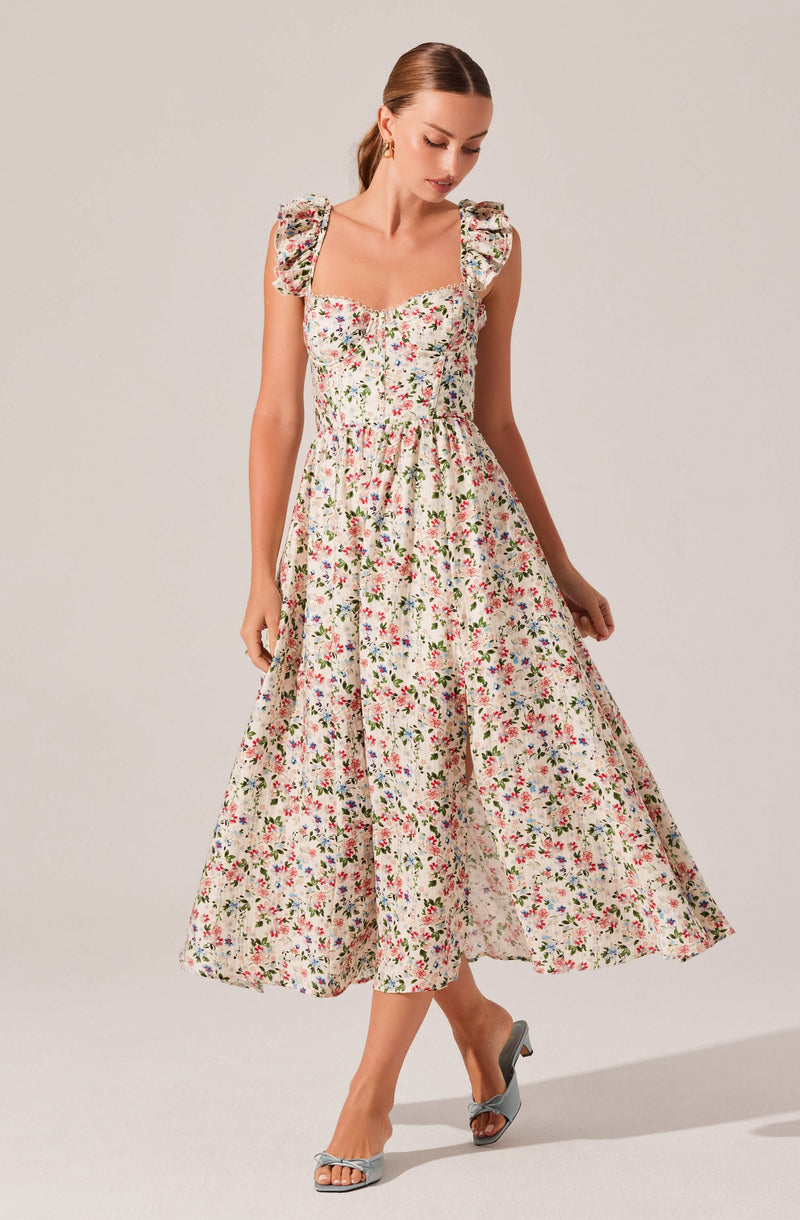 A woman is wearing a long, floral dress with a fitted bodice and ruffled shoulder straps. The dress has a white background adorned with small, colorful flowers in red, pink, blue, and green. The skirt of the dress is full and flows down to her ankles. She is also wearing light blue high-heeled sandals with bows on the front.