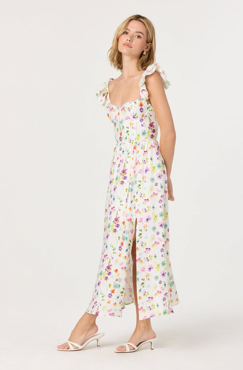 A woman is wearing a sleeveless, ankle-length dress with a floral print featuring pink, purple, orange, green, and yellow flowers on a white background. The dress has ruffled shoulder straps and a fitted bodice, with a front slit that reveals her legs. She is also wearing strappy, open-toed high-heeled sandals in a light color.