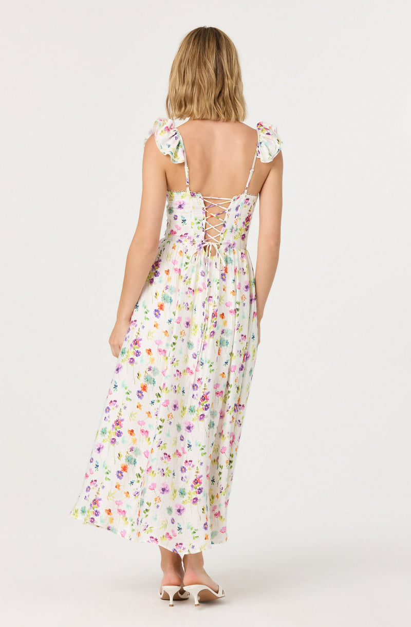 A woman is wearing a long, flowing dress with a colorful floral print on a white background. The dress features short ruffled sleeves and a lace-up back with crisscrossed laces. She is also wearing a pair of white high-heeled shoes.