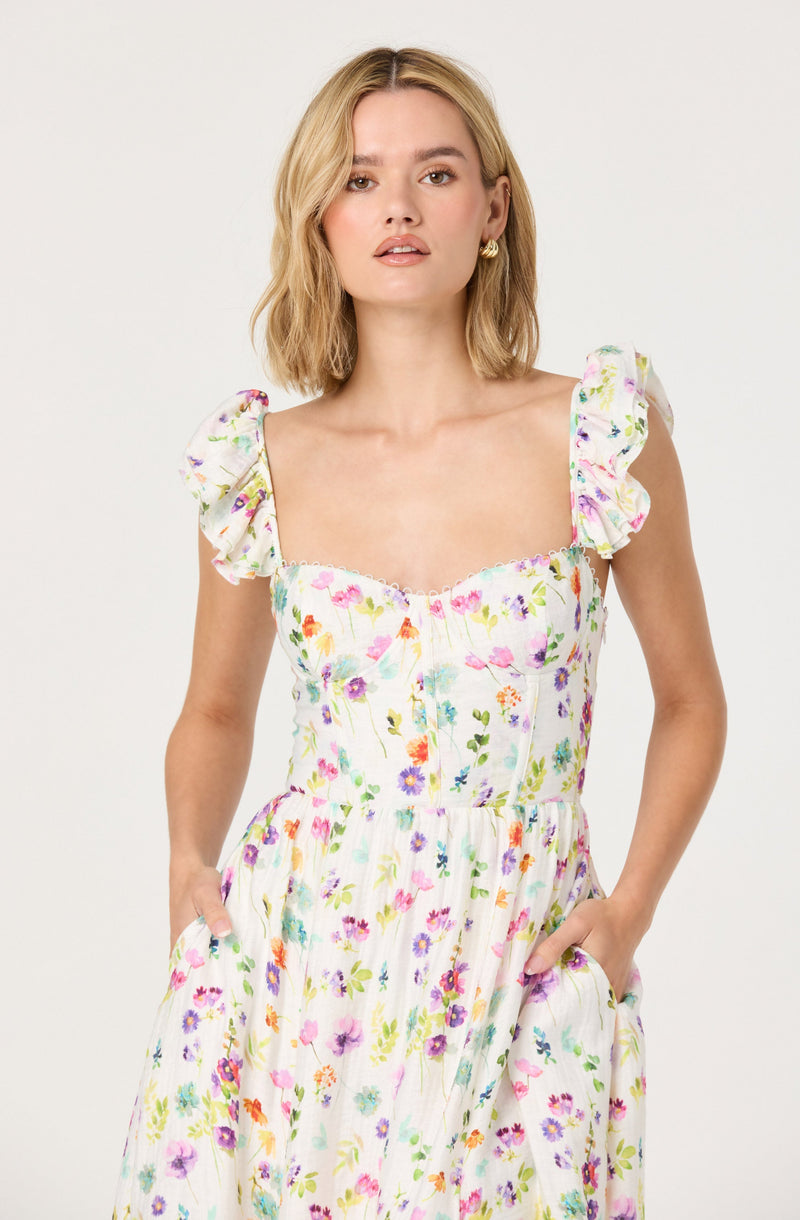 A woman wears a light, sleeveless dress with bright, colorful floral patterns. The dress has ruffled cap sleeves and a fitted bodice with vertical seaming and gathered fabric. She has her hands in the dress's side pockets.