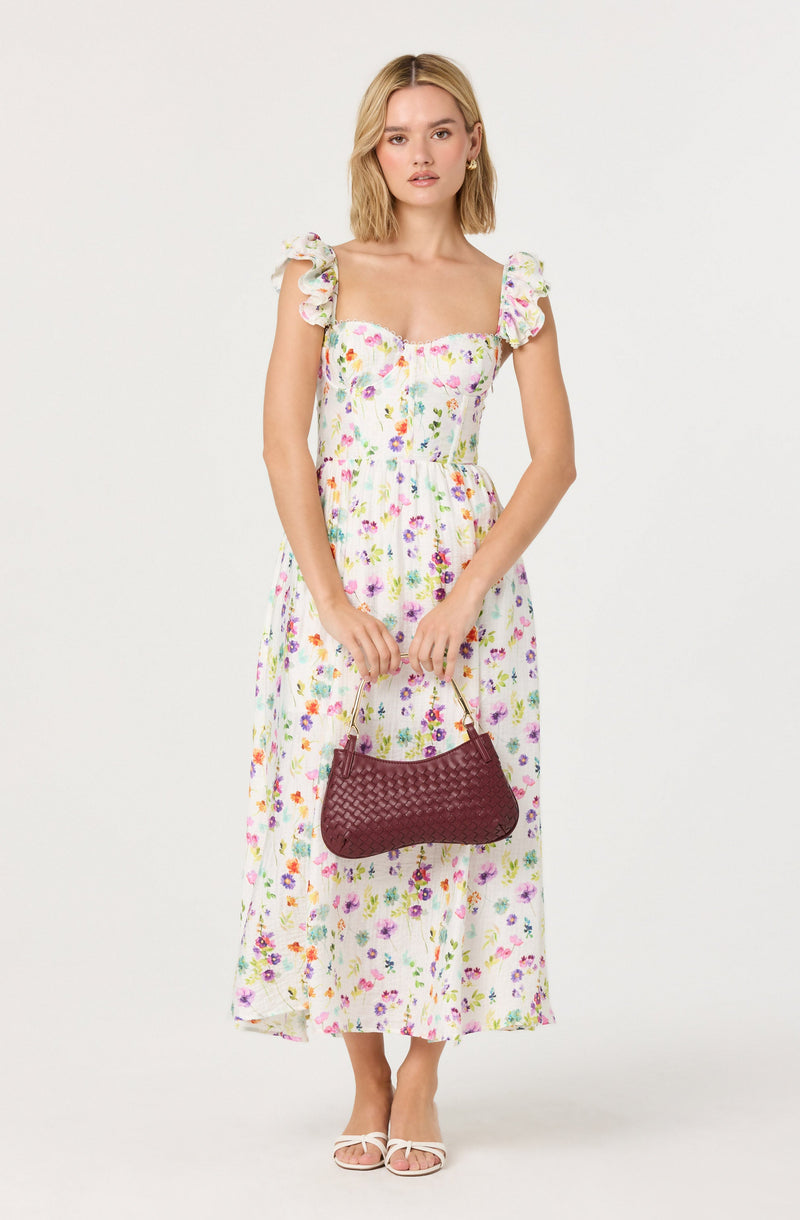 A woman is wearing a long, white dress with a colorful floral pattern. The dress has ruffled, puffed sleeves and a fitted bodice with a square neckline. She is holding a maroon, woven handbag with a gold handle and is wearing white, open-toed heeled sandals. She has shoulder-length blonde hair and is standing against a plain, light background.