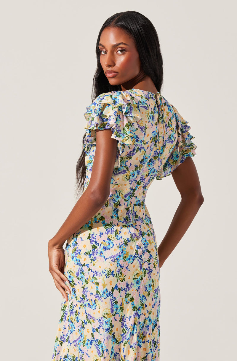 A woman is wearing a fitted dress with a floral print featuring shades of blue, purple, green, and yellow. The dress has ruffled short sleeves with multiple layers of ruffles, creating a voluminous look, and a high neckline. The fabric appears lightweight and has a smooth texture.
