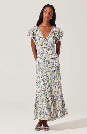 A woman is wearing a long, flowy dress with a floral pattern in shades of blue, purple, green, and beige on a light background. The dress features a V-neckline and short, ruffled sleeves.