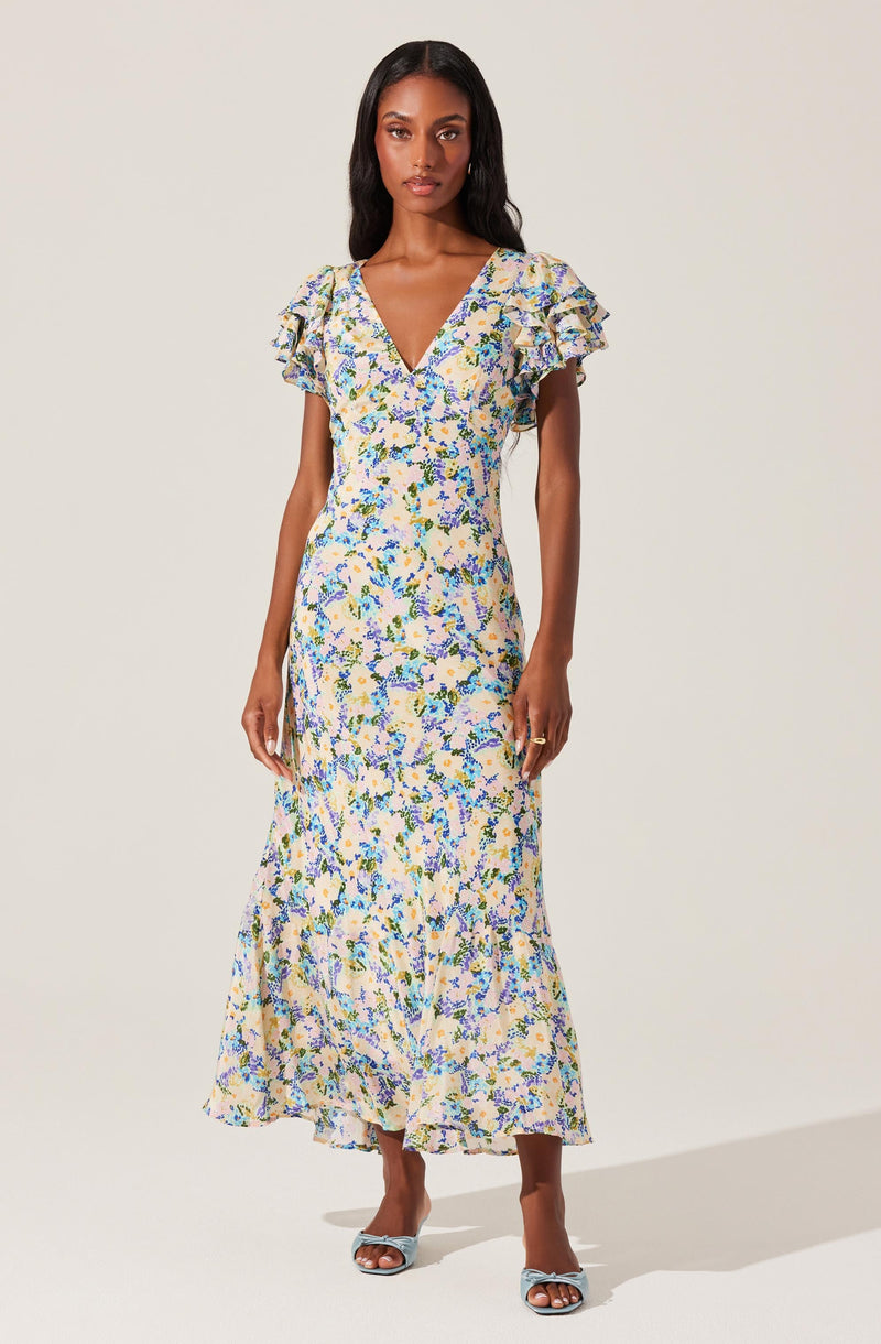 A woman is wearing a long, flowy dress with a floral pattern in shades of blue, yellow, and green on a light background. The dress features a V-neckline and short ruffled sleeves. She is also wearing a pair of open-toed heels with a bow detail.