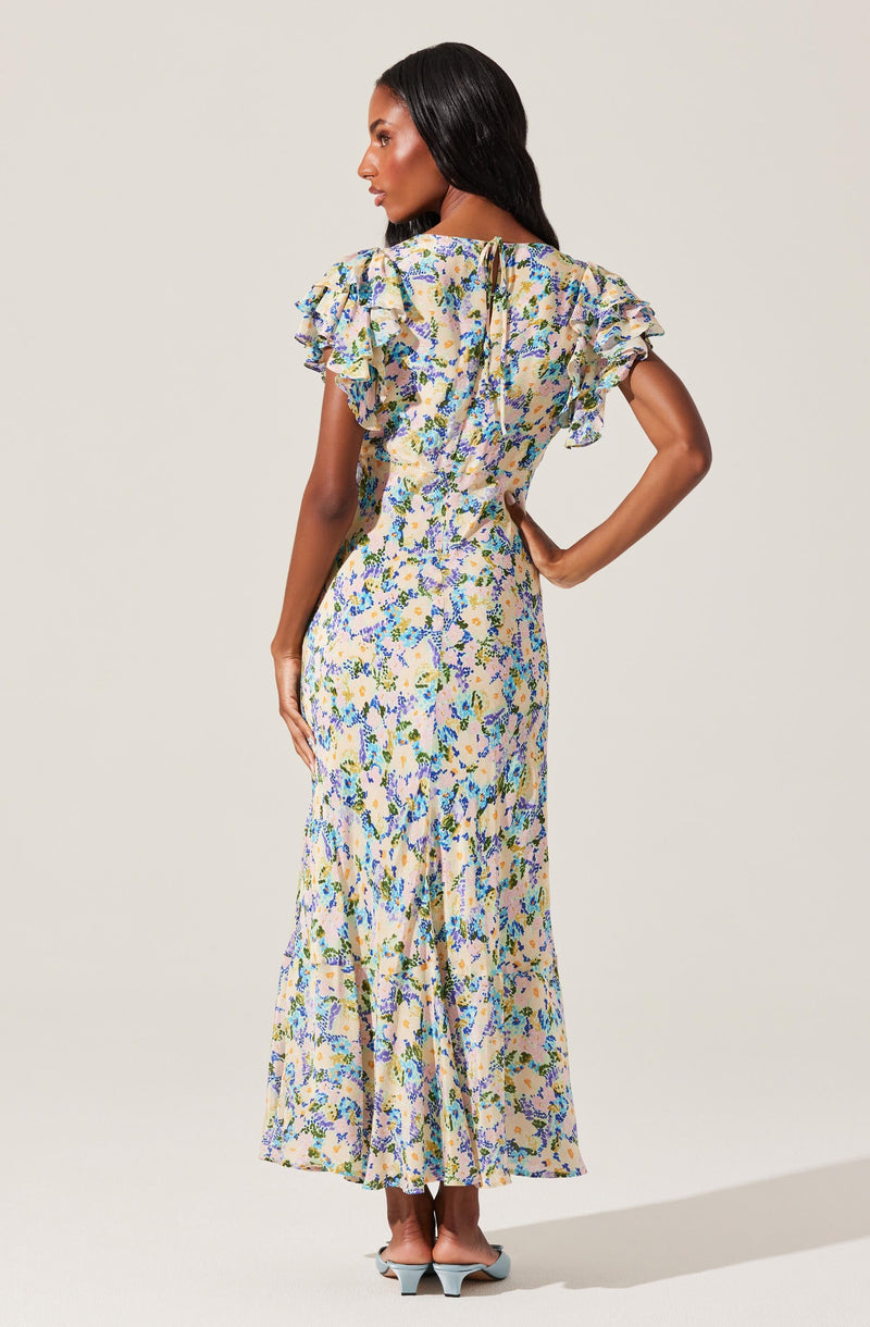 A woman is wearing a long, floral-patterned dress with ruffled short sleeves. The dress features a fitted waist and a flowing skirt that reaches the floor. The fabric has a light background with a multicolored floral design, primarily in shades of blue, green, yellow, and pink. She is also wearing light blue pointed-toe shoes with a low heel.