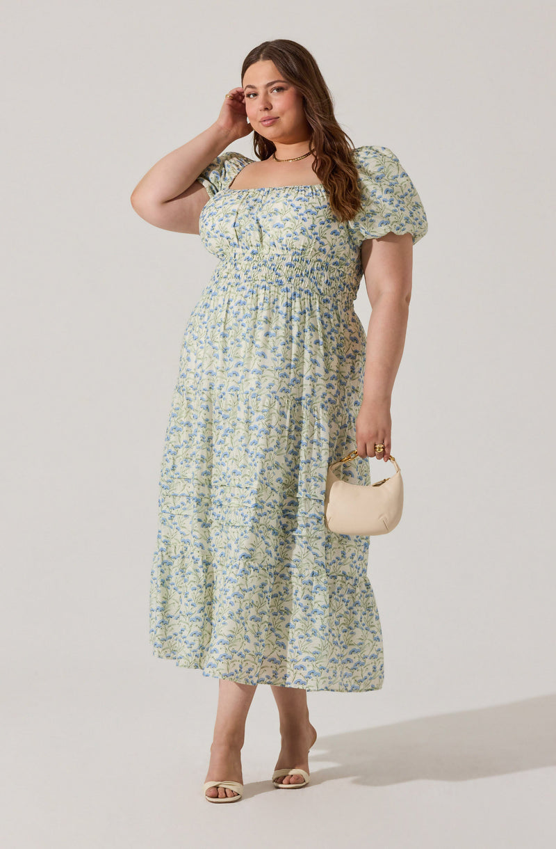 A woman is wearing a light-colored, floral-patterned dress with puffed short sleeves. The dress has a square neckline and is fitted at the waist with gathered fabric, flowing down to ankle length. She is holding a small, cream-colored handbag with a short handle and is wearing open-toed heels.
