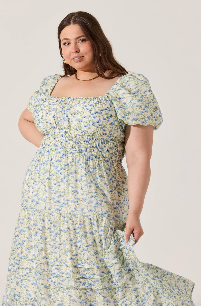 A woman is wearing a light-colored, floral dress with a square neckline and puffed short sleeves. The dress features a smocked waist and a flowing skirt with a similar floral pattern in shades of blue and green. She accessorizes with small earrings and a choker necklace.