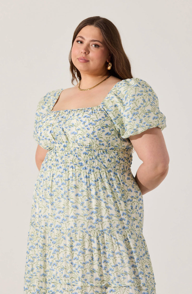 A woman is wearing a light-colored dress with a floral pattern of blue and green leaves and flowers. The dress has a square neckline, puffed short sleeves, and a smocked waist.