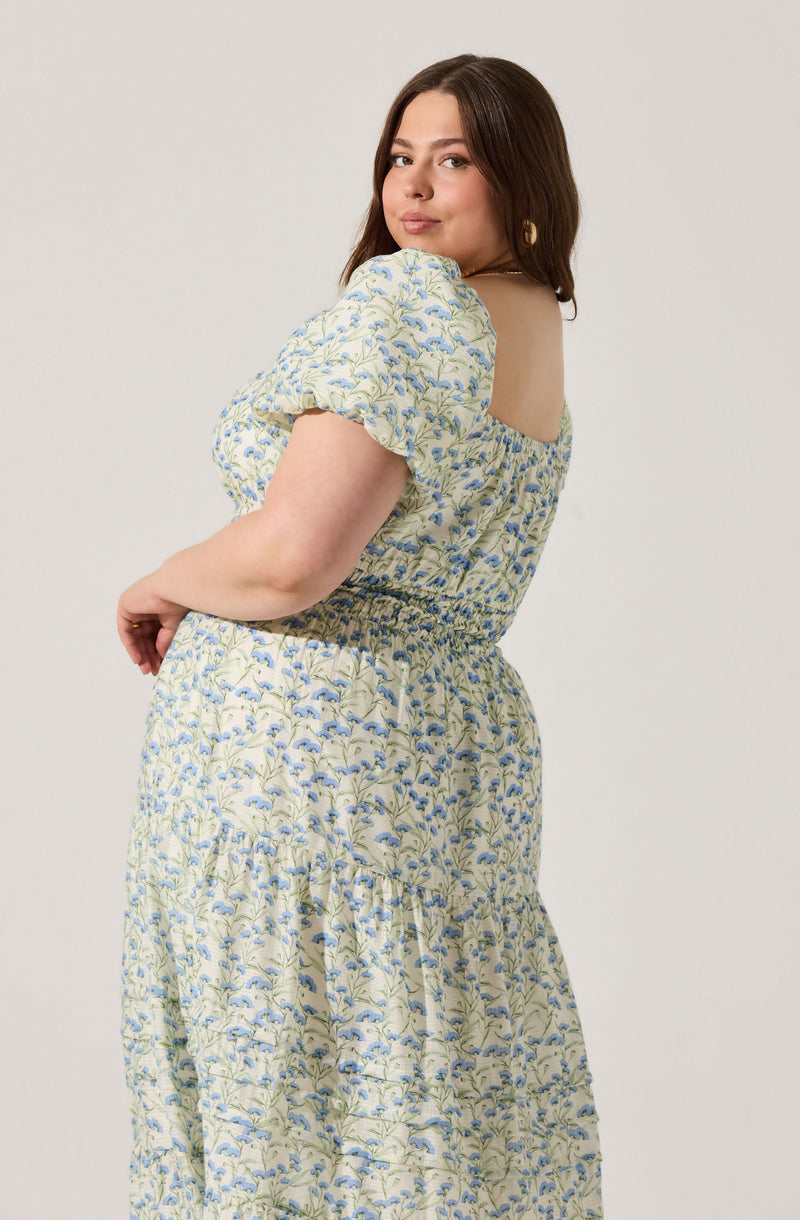 A woman is wearing a light-colored dress with a floral pattern featuring blue and green leaves and flowers. The dress has puffed short sleeves, an elasticized waist, and a tiered skirt.