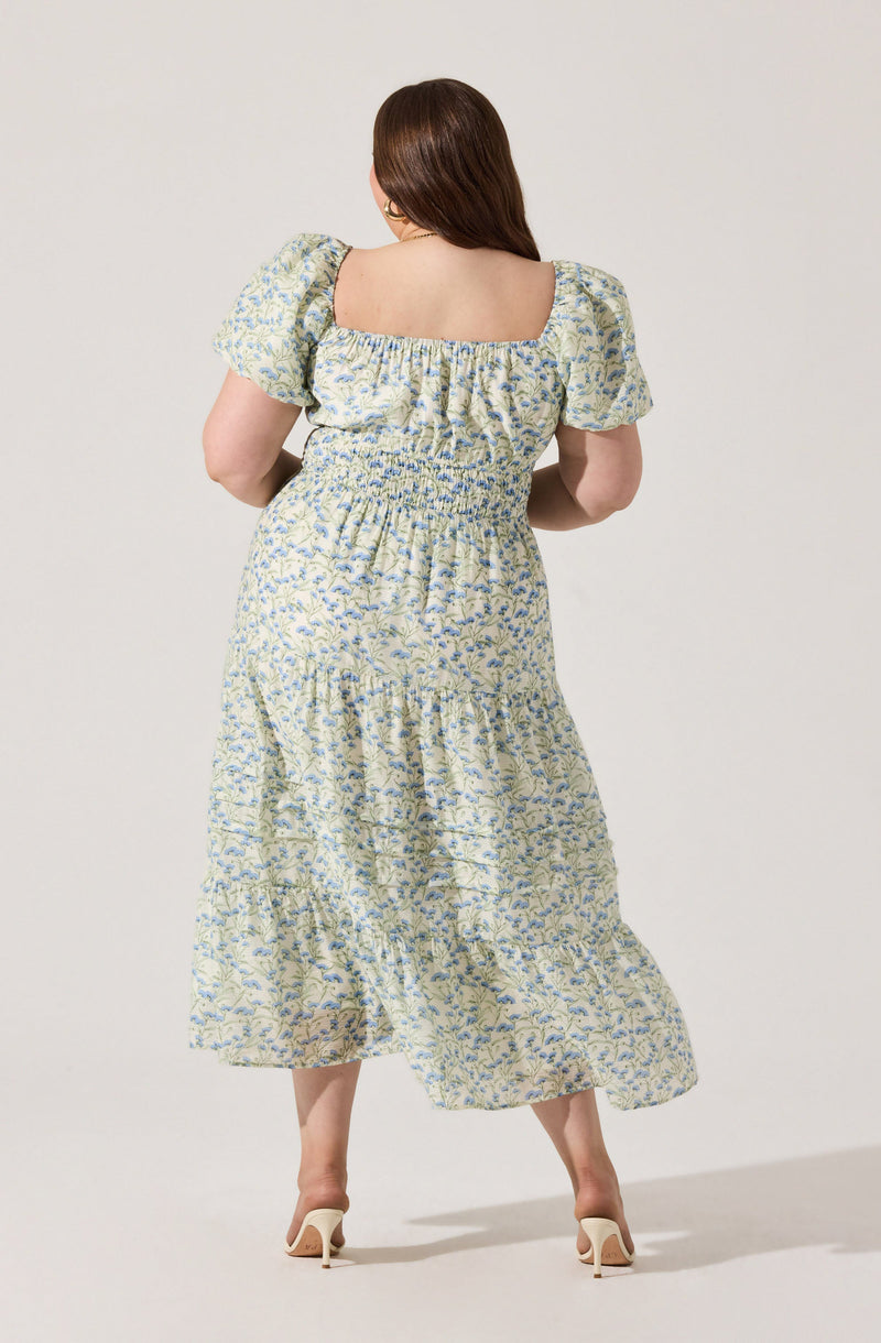 A woman is wearing a light-colored, floral-patterned dress with a square neckline and puffed short sleeves. The dress has a smocked waist and a tiered, flowing skirt that reaches mid-calf. She is also wearing white high-heeled shoes.
