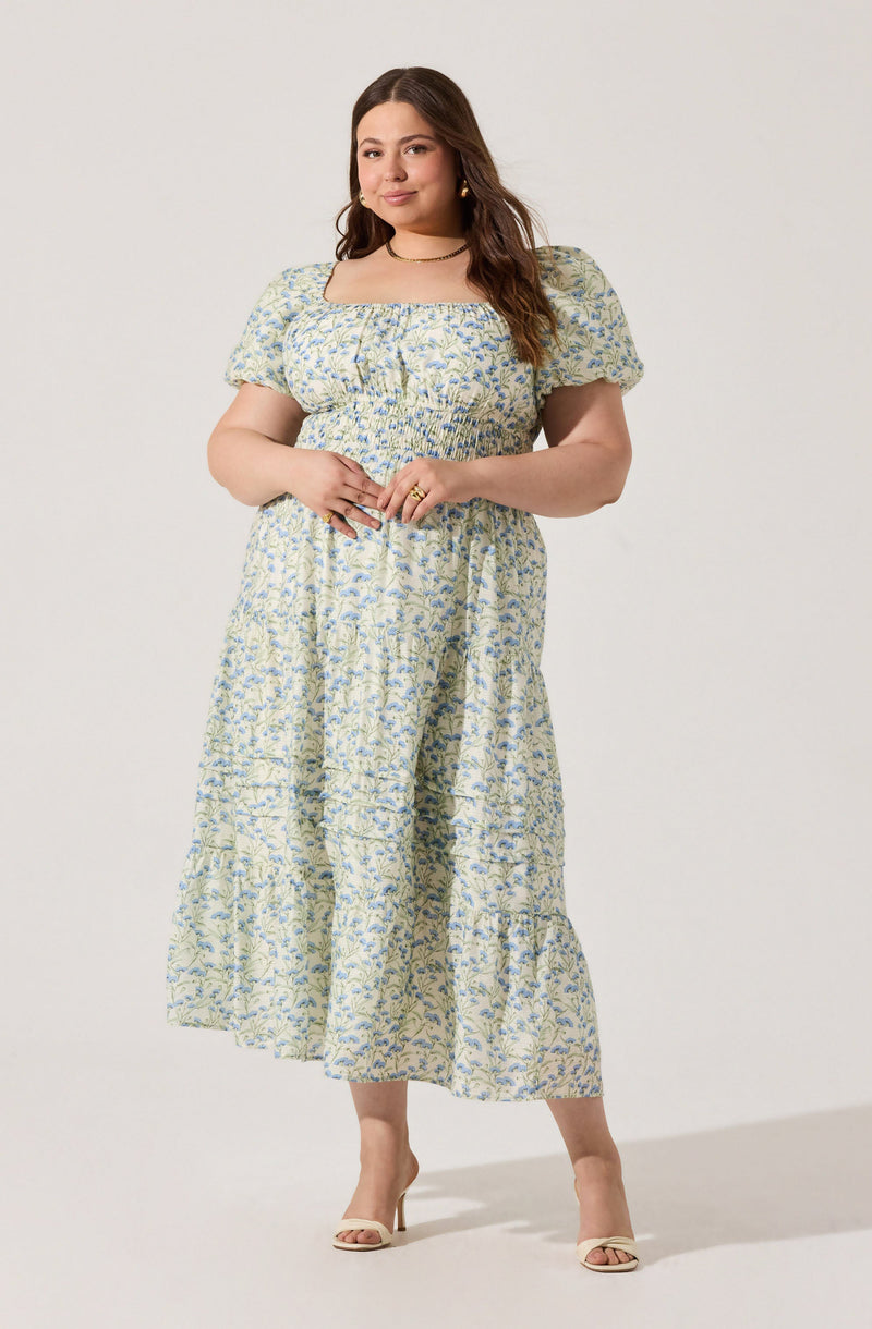 A woman is wearing a light-colored, floral-patterned dress with short puffed sleeves and a square neckline. The dress has a smocked waistband and a tiered, ankle-length skirt. She is also wearing beige open-toe heels, gold hoop earrings, and a delicate choker necklace.