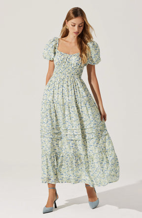 A woman is wearing a long, light-colored dress with a floral pattern. The dress features puffed short sleeves, a square neckline, and gathered fabric at the waist creating a slightly fitted bodice. The skirt is full and flowing, reaching down to her ankles. She is also wearing pointed-toe heels in a light gray color with black heels.