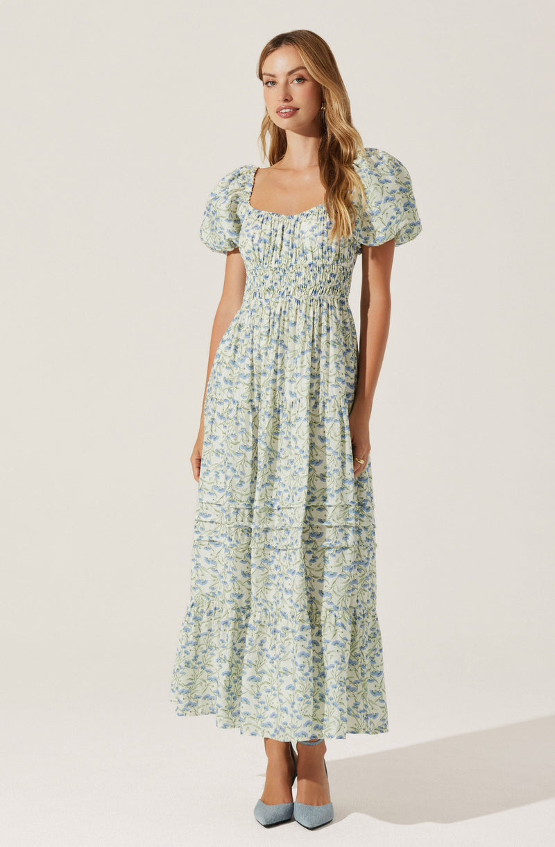 A woman is wearing a long, flowing dress with a floral pattern in shades of blue and green on a light-colored background. The dress features a square neckline, puffed short sleeves, and a fitted bodice with ruching detail. The skirt portion is gathered at the waist and extends down to the ankles.