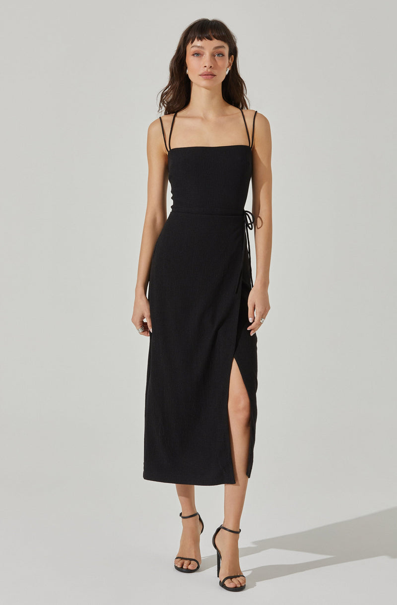 A woman is wearing a black sleeveless dress with thin spaghetti straps. The dress has a straight neckline, a side tie detail, a slit on one side revealing her leg, and a textured fabric. She is also wearing black strappy high heel sandals.