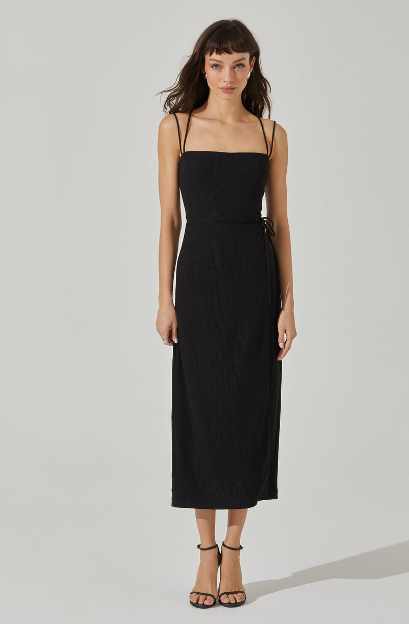 A woman is wearing a long, black, sleeveless dress with spaghetti straps. The dress has a fitted bodice and a wrap-around style with a side tie. She is also wearing black high-heeled sandals with ankle straps.