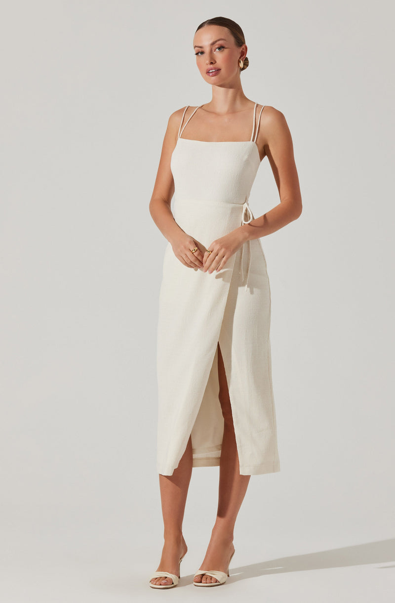 A woman is wearing a sleeveless, cream-colored dress with thin spaghetti straps. The dress has a fitted bodice and a long skirt with a front slit, allowing a glimpse of her legs. She is accessorizing with gold earrings and rings, and she is wearing neutral-colored high-heeled sandals.