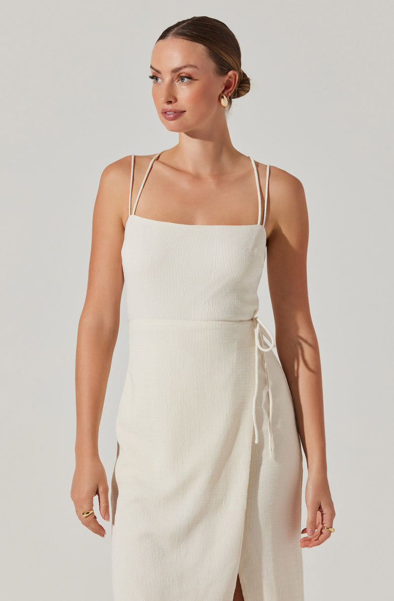 A woman is wearing a sleeveless, off-white ribbed dress with thin spaghetti straps that cross at the shoulders. The dress is fitted at the top and features a side tie detail. The fabric appears textured, and the overall look is elegant and minimalist. She has her hair styled in a low bun and accessorizes with gold hoop earrings and a gold ring on her finger.
