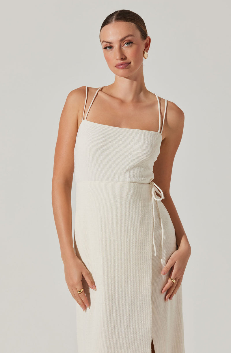 A woman is wearing a sleeveless, cream-colored dress with thin, double shoulder straps. The fabric has a textured, ribbed pattern, and the dress features a side tie detail at the waist.