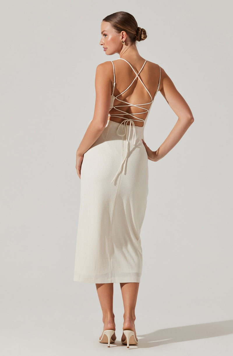 A woman is wearing a backless, cream-colored dress with thin, crisscross straps on the upper part that tie around her waist. She is also wearing nude high-heeled shoes and gold hoop earrings.