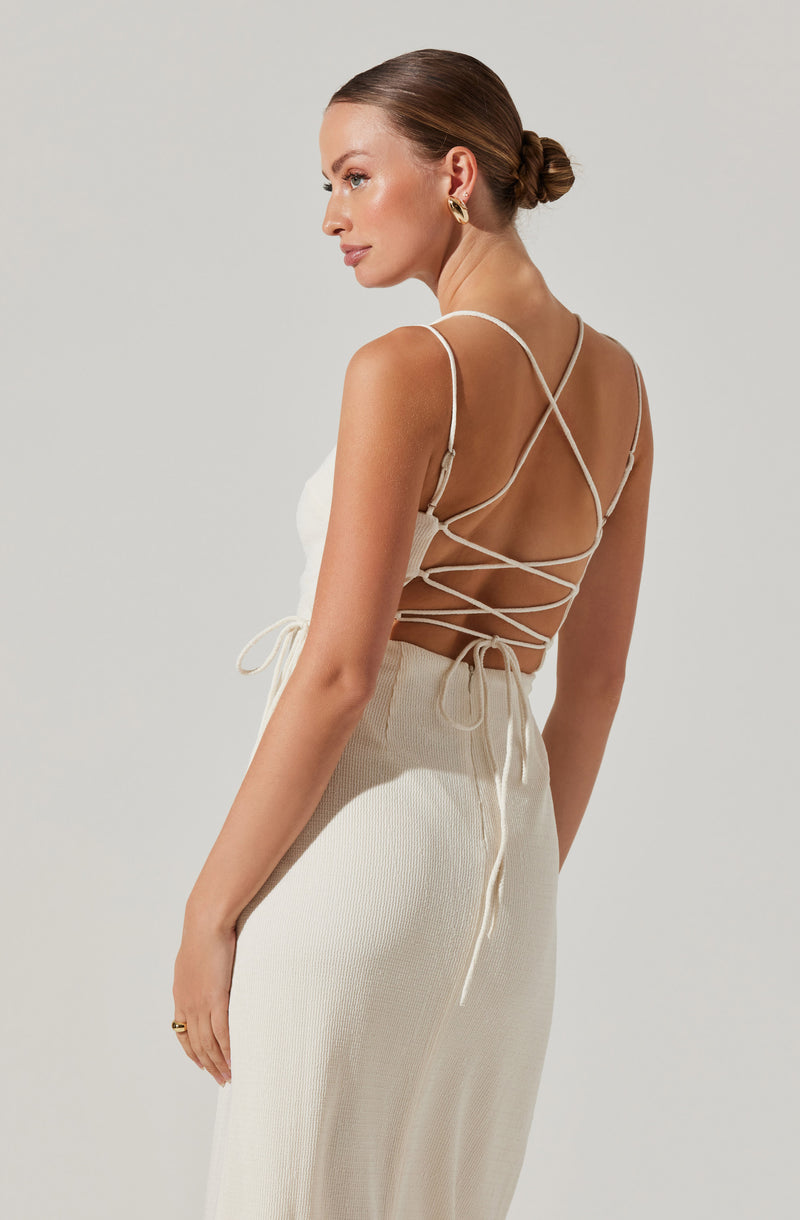 A woman is wearing a sleeveless, backless cream-colored top with delicate spaghetti straps crossing and tying at the back. She is also dressed in high-waisted, cream-colored knit pants that have a textured appearance.
