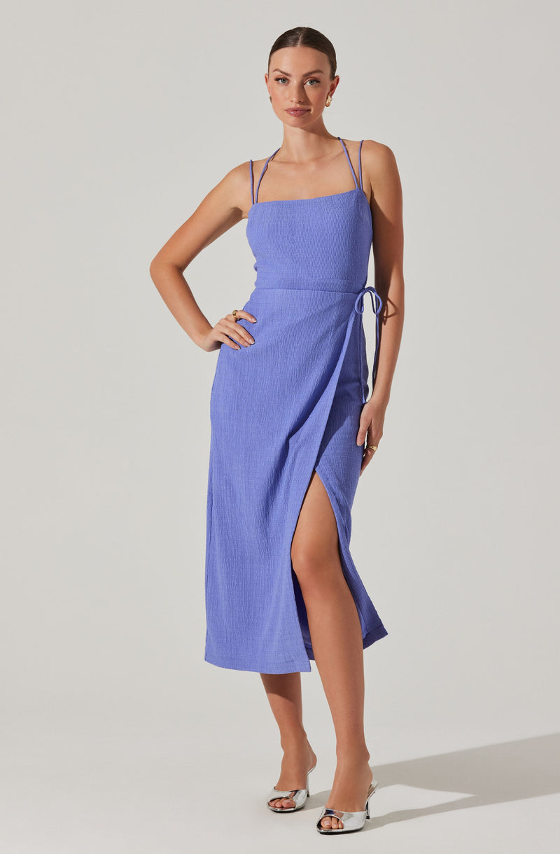 A woman is wearing a sleeveless, ankle-length dress in a textured, light purple fabric. The dress features thin spaghetti straps, a fitted bodice, and a side slit that reveals part of her leg. It has a drawstring detail at the waist. She is also wearing open-toe heels with a metallic finish.