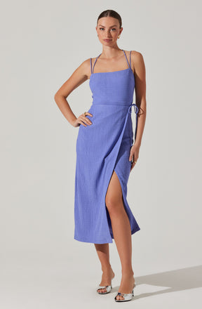 A woman is wearing a sleeveless, ankle-length dress in a textured, light purple fabric. The dress features thin spaghetti straps, a fitted bodice, and a side slit that reveals part of her leg. It has a drawstring detail at the waist. She is also wearing open-toe heels with a metallic finish.