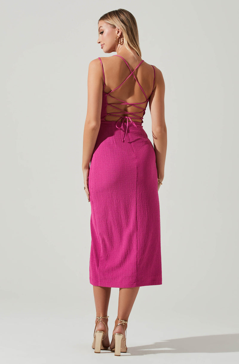 A woman is wearing a sleeveless, pink, knit dress with spaghetti straps. The back of the dress features crisscrossing spaghetti straps and laces that tie at the waist, creating a revealing open-back design. She has on beige high-heeled sandals and gold hoop earrings.