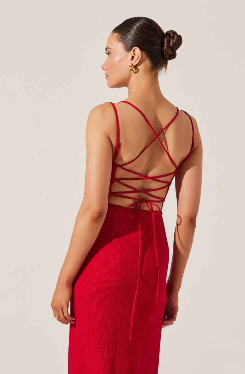 A woman is wearing a sleeveless red dress with thin straps over her shoulders. The back of the dress features an intricate design with multiple crisscrossing straps and a tie at the lower back. The dress extends to a long skirt made of a textured fabric. She has her hair styled in a sleek bun and accessorizes with large gold hoop earrings.