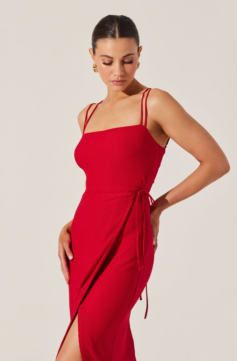 A woman is wearing a fitted, ribbed, red dress with thin double straps over her shoulders. The dress features a side tie detail at the waist and a slit along the front.